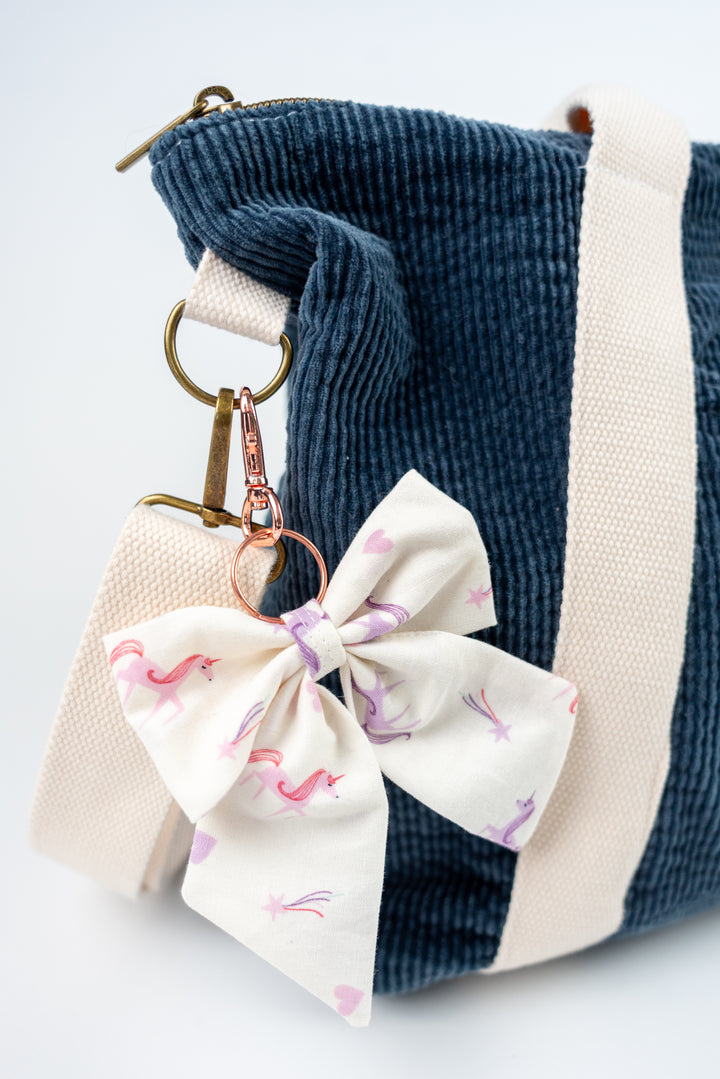 Keychain with a white bow featuring unicorns attached to a blue corduroy bag.