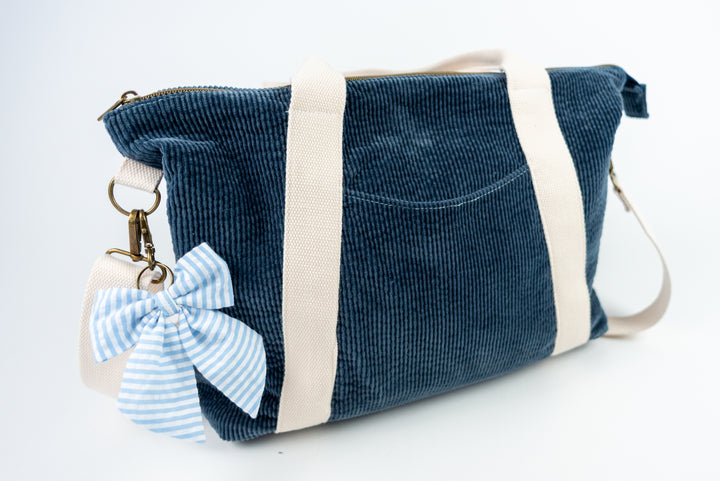 Blue corduroy bag with a white strap and a blue and white striped bow keychain on a white background