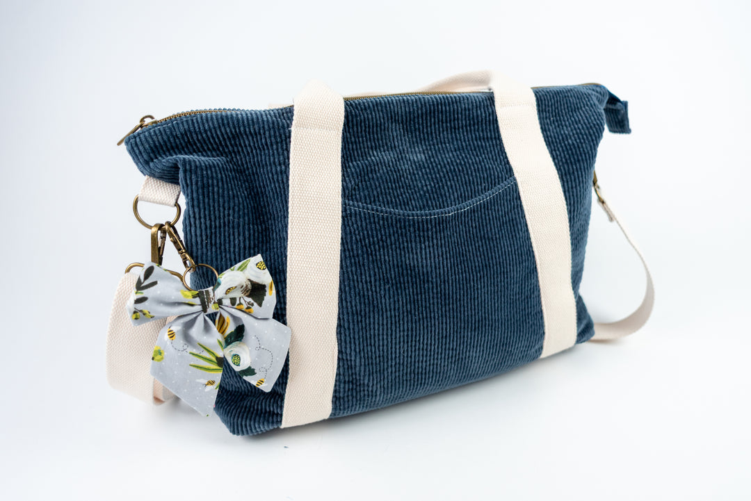 Blue corduroy bag with a grey floral bow charm on a white background