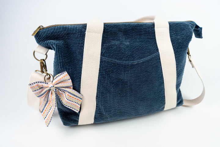 Blue corduroy bag with a bow keychain that is pink striped with multiple colors on a white background
