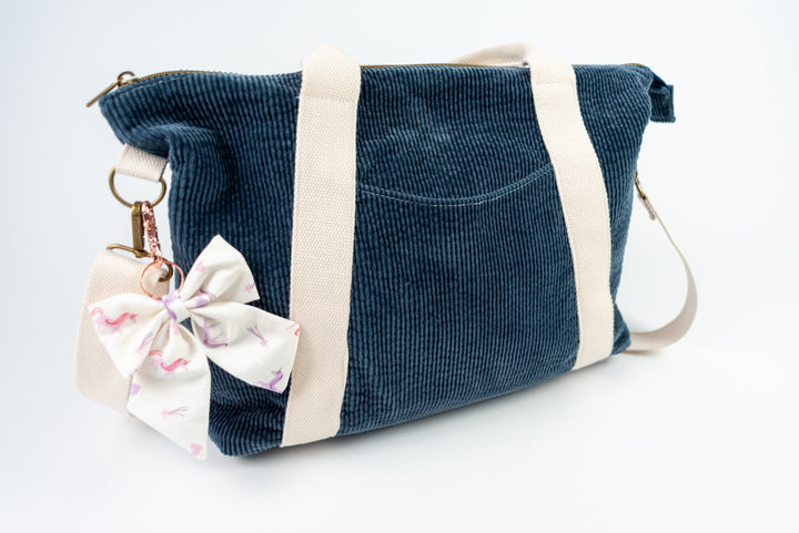 Blue corduroy bag with a white strap and a bow keychain on a white background